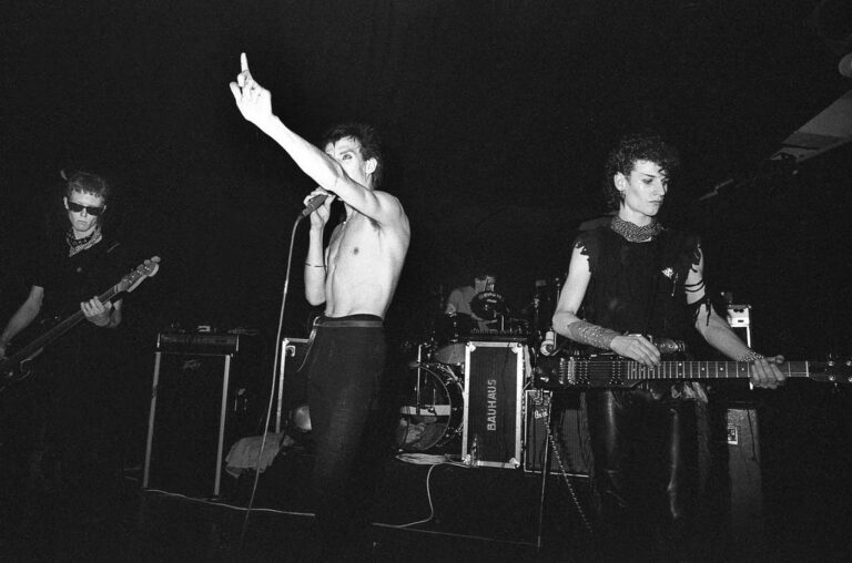 Black and white photo of iconic gothic rock band Bauhaus performing live on stage, with shirtless goth singer holding a microphone and raising his middle finger, surrounded by band members playing dark wave and post-punk music, representing classic 80s goth bands and pioneers of gothic rock music.