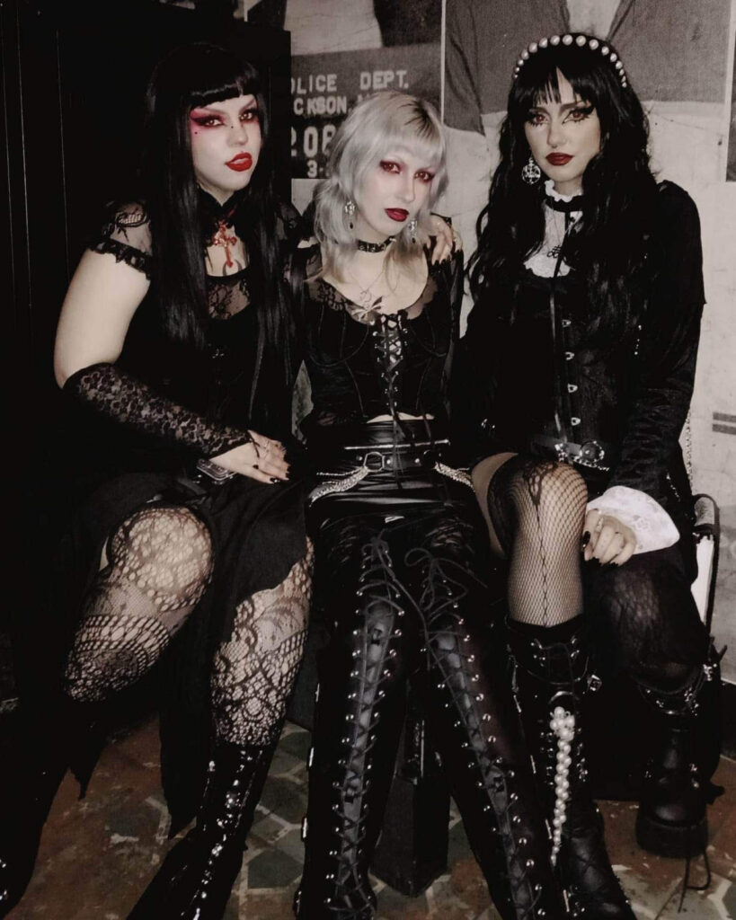 Three gothic women in black lace and leather outfits posing in dark club setting, wearing dramatic makeup and platform boots, representing modern goth fashion and style influenced by gothic rock, goth punk, and emo goth aesthetics, reflecting contemporary goth bands and gothic subculture.